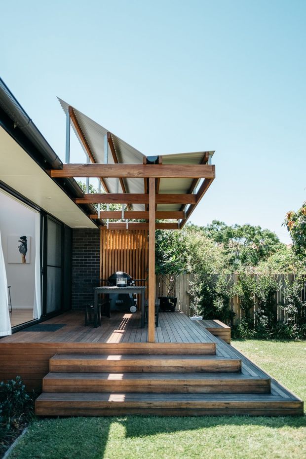 A residential outdoor timber deck and entertaining area adjoining a contemporary house. Broad timber steps lead to a raised deck with a built-in-style timber pergola, exposed structural beams, translucent roof panels, vertical timber screening, and a barbecue. The space opens onto a landscaped backyard and is photographed in bright daylight.