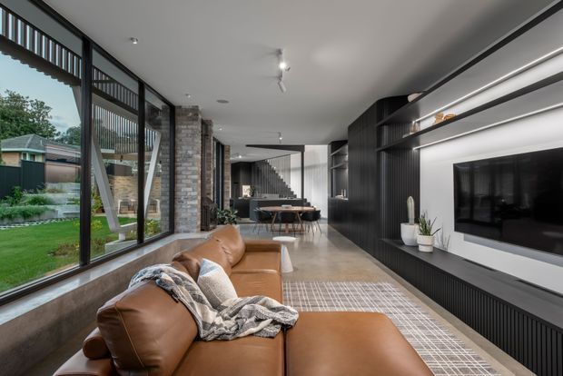 A contemporary open-plan living room with a large brown leather sectional sofa, integrated media wall and television, dark vertical-panel cabinetry, open shelving, exposed grey brick columns, polished concrete flooring, and extensive floor-to-ceiling glazing. The space extends toward a dining area and staircase, with a suburban garden visible outside.