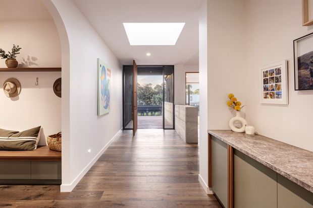 A bright, contemporary residential hallway and entry zone with warm timber flooring, white walls, and a large skylight. The corridor leads to glazed doors opening onto an exterior balcony or terrace. On the left is a built-in arched display and seating nook with timber shelving, while the right side features a long stone-topped built-in cabinet or console with framed artwork and decorative objects.