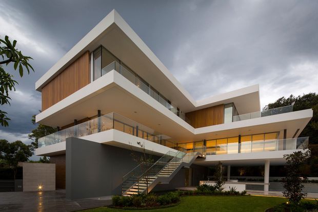 Exterior architectural photograph of a large contemporary multi-level residence with dramatic cantilevered white floor slabs, extensive glazing, glass balustrades, warm vertical timber wall cladding, and an exposed external staircase. The home sits in a landscaped suburban setting beneath a dark overcast sky.