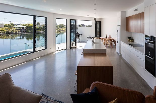 Bright open-plan contemporary kitchen and living area overlooking a calm waterfront through expansive black-framed glazing. The space features polished concrete flooring, white cabinetry with warm timber accents, a large island with a pale stone benchtop, integrated appliances, minimalist pendant lighting, and a dining area opening onto an outdoor terrace.