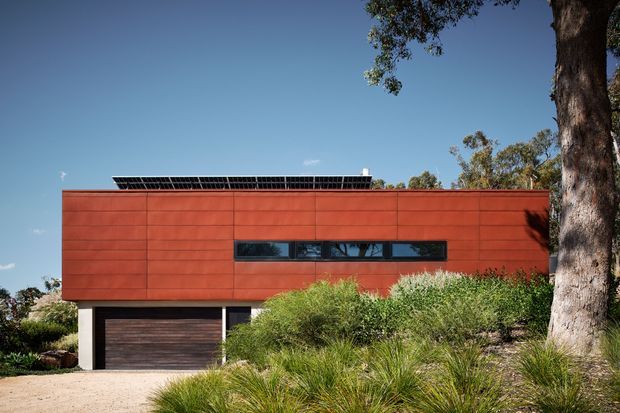 Contemporary single-storey residential facade with a long horizontal form, clad in warm red-brown panels and punctuated by a narrow horizontal strip window. A dark timber-toned garage door sits beneath the main volume, while native grasses and mature trees landscape the foreground. Solar panels are visible along the low roofline.