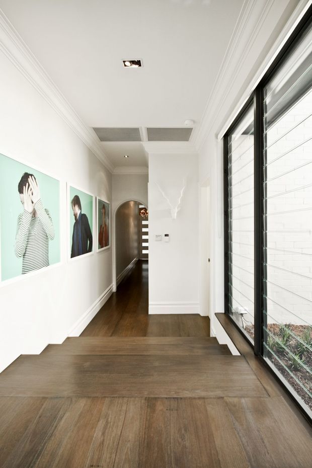A bright, refined residential hallway and stair landing with wide dark timber flooring, white walls, layered decorative cornices, recessed lighting, and framed artwork. A large black-framed glazed opening runs along the right side, overlooking a narrow planted courtyard or lightwell. The hallway continues through an arched opening toward the rear.