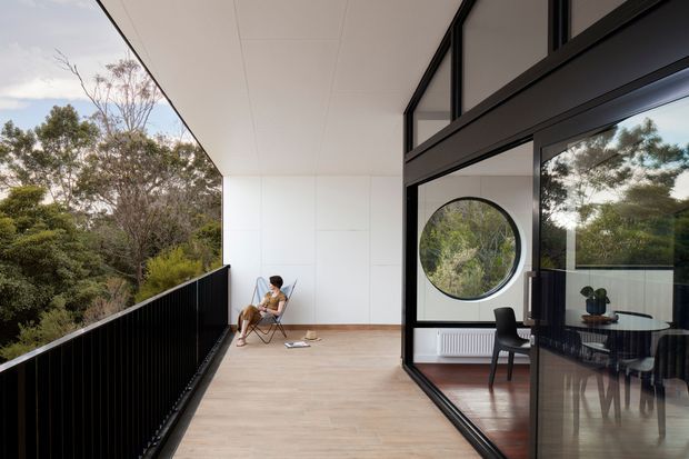 A contemporary elevated balcony or terrace adjoining a glazed living and dining area. The space features pale timber-look decking, a black metal balustrade, white wall and soffit panels, and expansive black-framed glazing with a distinctive circular window. Reflections and views reveal a furnished interior with a dining table and chairs, while surrounding trees create a leafy setting.