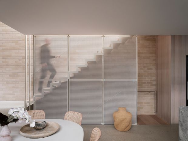 Contemporary interior view centered on a stairway enclosed behind translucent ribbed glass or polycarbonate screens with slim metal framing. Pale exposed brick walls, a smooth light ceiling, neutral flooring, and a partially visible dining area with a round table and woven chairs create a soft, understated palette. A blurred person is visible on the stairs behind the screening, with a timber-toned doorway or wall panel to the right.