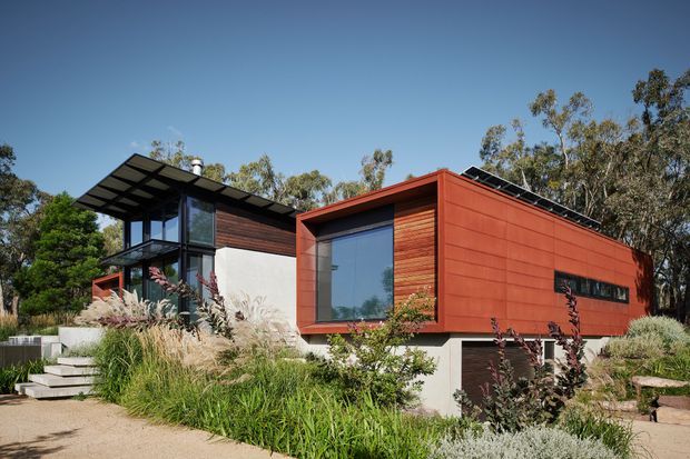 Exterior view of a contemporary multi-level house set within a landscaped garden. The building combines a rust-red metal-clad rectangular volume with dark timber wall cladding, extensive glazing, a projecting flat canopy, and a partially recessed lower level. Native-style planting, concrete steps, and a paved path surround the entrance.