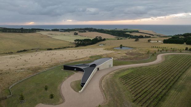 Aerial view of a sculptural low-profile building set within expansive rolling farmland near the coast. The building has a curved, dark roof and angular pale walls, surrounded by a driveway, cultivated rows, open fields, scattered trees, and a distant ocean horizon beneath an overcast sky.