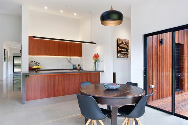 Bright contemporary open-plan kitchen and dining area with a warm timber island and wall cabinetry, white walls, polished concrete flooring, and a round dark wood dining table with sculptural chairs. Black-framed full-height glazing opens to a timber-clad outdoor area, while a large pendant light and linear kitchen lighting add visual interest.