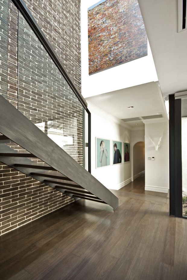 Bright contemporary residential interior showing a dramatic open-riser staircase with dark metal structure and timber treads against an exposed dark brick feature wall. The staircase rises beside a tall glazed opening, leading into a white hallway with timber flooring, framed artwork, recessed lighting, and a double-height void with a large abstract artwork.