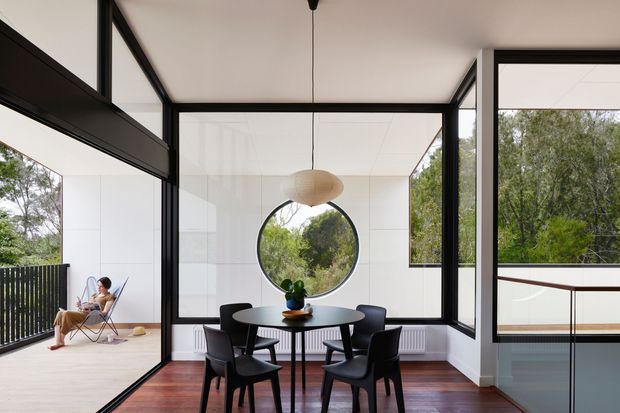 A bright contemporary dining area with a round black dining table and sculptural pendant light, enclosed by expansive black-framed glazing. The room opens to a balcony or terrace with outdoor seating, while mature greenery is visible beyond. Dark timber flooring, white walls and ceiling, and a circular feature window create a clean, modern composition.