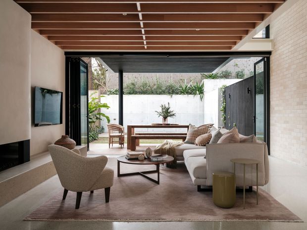 A refined contemporary living room opening directly onto a private landscaped courtyard through wide black-framed glazed doors. The room features a cream sectional sofa, sculptural armchair, layered round coffee tables, neutral rug, built-in fireplace and wall-mounted television. Warm timber ceiling battens contrast with pale plaster and light brick walls, while a timber dining table and courtyard planting are visible beyond.