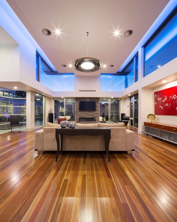A spacious, highly polished living room with a dramatic double-height void and extensive floor-to-ceiling glazing. A large beige sectional sofa faces a wall-mounted television and stone feature wall, with a dark console table in the foreground. Warm striped timber flooring contrasts with white walls, blue illuminated glazing, recessed spotlights, and a circular sculptural ceiling light. A red decorative artwork and low timber media cabinet add colour along the right wall.