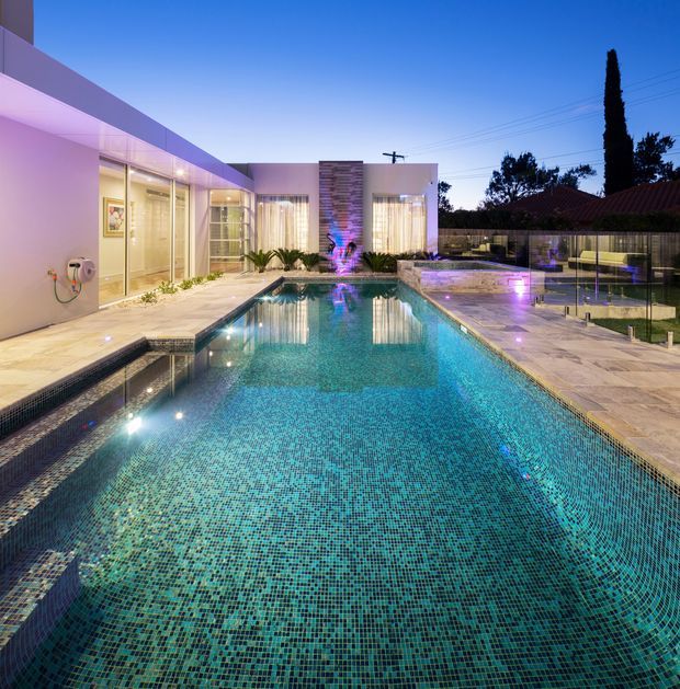 Twilight view of a contemporary residential pool courtyard. A large rectangular swimming pool with turquoise mosaic tiling and illuminated water dominates the foreground, bordered by pale stone paving and glass pool fencing. The surrounding single-storey home has crisp flat-roofed forms, pale rendered walls, extensive glazing, and subtle purple architectural lighting. A neighbouring tiled roof and mature trees are visible beyond the property.