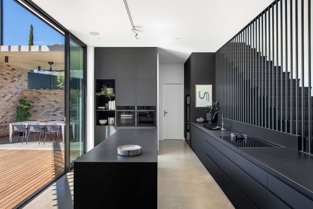 A contemporary black-and-white kitchen with a long island and extensive dark built-in cabinetry. Integrated ovens and appliances are set into the rear cabinetry, while a linear kitchen run with cooktop and sink occupies the right wall beneath a striking vertical black slat screen. Full-height glass doors open to a timber deck and outdoor dining area, bringing in abundant natural light. The space has polished concrete flooring, minimalist detailing, and a refined monochrome palette.