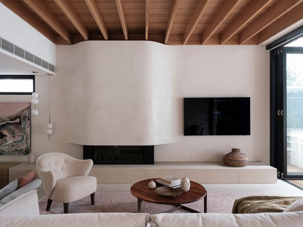 Warm, refined contemporary living room with a broad plastered fireplace and media wall, built-in hearth bench, sculptural lounge chairs, a round timber coffee table, and a large upholstered sofa. Exposed timber ceiling beams and decking create a strong architectural feature, while full-height black-framed glazing opens to the exterior.