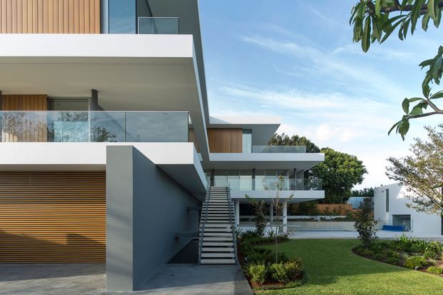 Exterior architectural view of a contemporary multi-level residence with crisp white cantilevered slabs, dark grey rendered walls, extensive clear glass balustrades, vertical timber screening and horizontal timber garage detailing. An external stair connects the landscaped lawn and lower levels, with a neighboring modern wing and garden visible in the background.
