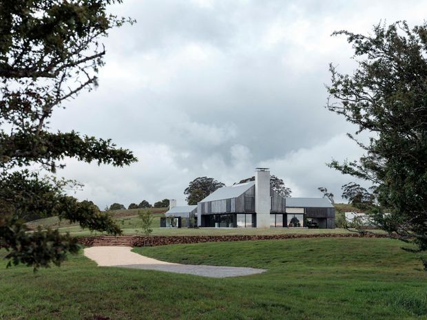 A contemporary single-storey rural residence set within expansive landscaped grounds. The house has a long, low horizontal form with dark glazed and clad wings, a pale central masonry chimney or vertical core, and a shallow pitched metal roof. A curved paved walkway leads through the lawn toward the home, with low stone retaining walls and mature trees framing the view beneath an overcast sky.