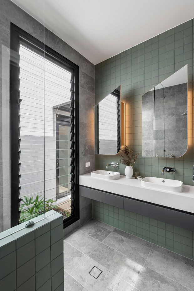 Contemporary bathroom with a double vanity, two white vessel basins, wall-mounted dark cabinetry, muted green square wall tiles, and large grey stone-look floor and wall tiles. A black-framed louvred window provides natural light, while rounded-edge mirrors and warm integrated lighting add a refined finish.