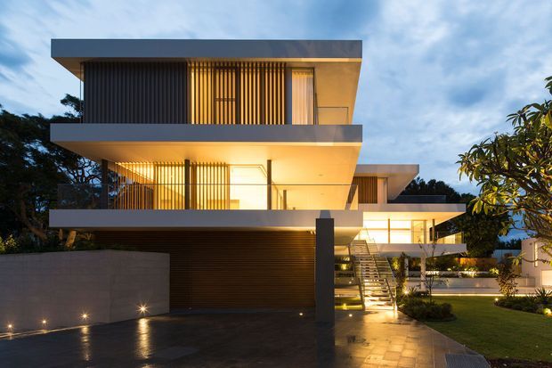 Exterior architectural photograph of a contemporary multi-level residence at dusk. The house features strong horizontal slab roofs and balconies, floor-to-ceiling glazing, vertical timber or metal screening, warm integrated lighting, a dark garage door, a paved driveway, landscaped lawn, and an external stair leading toward a recessed entry.