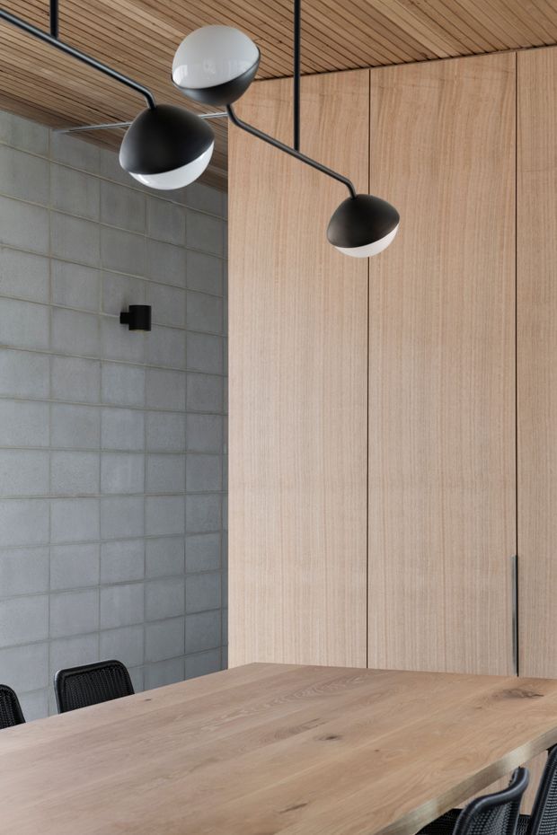 A refined dining area with a large light timber dining table and black woven chairs. The room features floor-to-ceiling light timber cabinetry or wall panelling, a grey concrete-block wall, a matching timber slatted ceiling, and sculptural black-and-white pendant lights on exposed black conduits. The palette is warm timber, grey, black, and white with a contemporary architectural character.