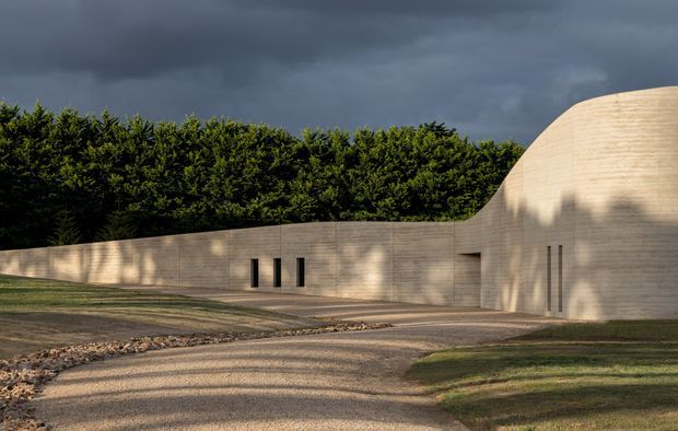 Exterior view of a sculptural, low-profile building integrated into a landscaped site. Long curved pale masonry or rendered walls form a continuous facade, with narrow vertical slit windows and a rounded roofline or vaulted volume. A gravel driveway and clipped hedges frame the composition beneath dramatic overcast skies.