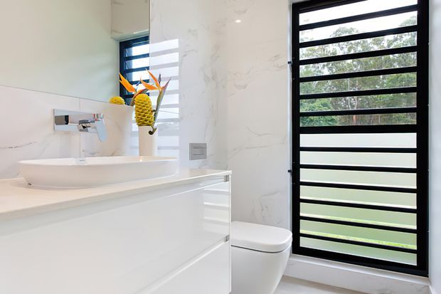 Bright contemporary bathroom with a white floating vanity, oval vessel basin, wall-mounted chrome tapware, white marble-look wall finishes, concealed storage, and a white toilet. A large black-framed louvred window provides strong contrast and views to greenery outside. Decorative tropical flowers are reflected in the mirror.