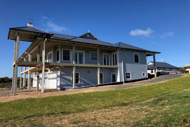 Exterior view of a large raised two-storey house with grey horizontal weatherboard cladding, a dark metal roof, broad covered verandas, balconies, exposed posts, and an open lower level. The home sits on a gently sloping grassed site under a clear blue sky, with neighbouring buildings visible in the background.