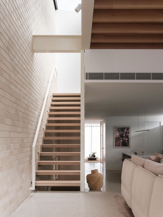 Interior view of a refined contemporary home showing an open timber staircase beside a pale exposed brick wall. The stair rises through a bright vertical void with skylight-like daylight, while the adjacent living area contains a pale upholstered sofa, artwork, a slim pendant light and timber ceiling lining. The palette is warm neutral with natural timber and light masonry finishes.