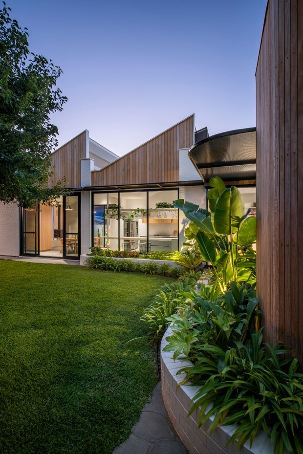 A contemporary residential courtyard viewed from the landscaped garden, featuring a single-storey pavilion-like house with warm vertical timber cladding, extensive black-framed glazing, and a folded pitched roofline. The transparent living spaces open onto a lush lawn and tropical planting, with a curved brick-edged garden bed and a distinctive curved metal canopy over the glazed corner.