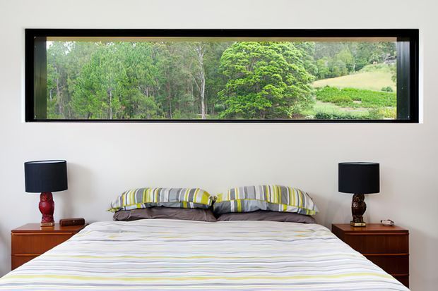 A contemporary bedroom with a low-profile bed dressed in striped white, grey, and lime-green bedding, flanked by matching dark timber bedside tables and black-shaded lamps. A long horizontal black-framed picture window provides a broad view of lush trees and rolling countryside.
