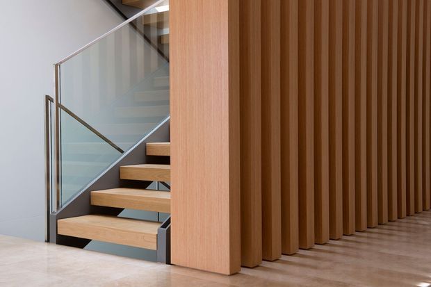 Contemporary interior stairway with floating timber treads, a clear glass balustrade, and a sculptural vertical timber screen forming a partial partition. The space has a restrained, refined palette of warm wood, pale flooring, and light neutral walls.