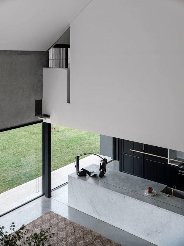 A contemporary double-height kitchen and living area with a large pale stone island, dark built-in cabinetry, polished concrete flooring, expansive floor-to-ceiling glazing, and a sculptural stair or mezzanine railing. The minimalist interior overlooks a lawn and uses strong black, grey, and white contrasts with clean geometric forms.