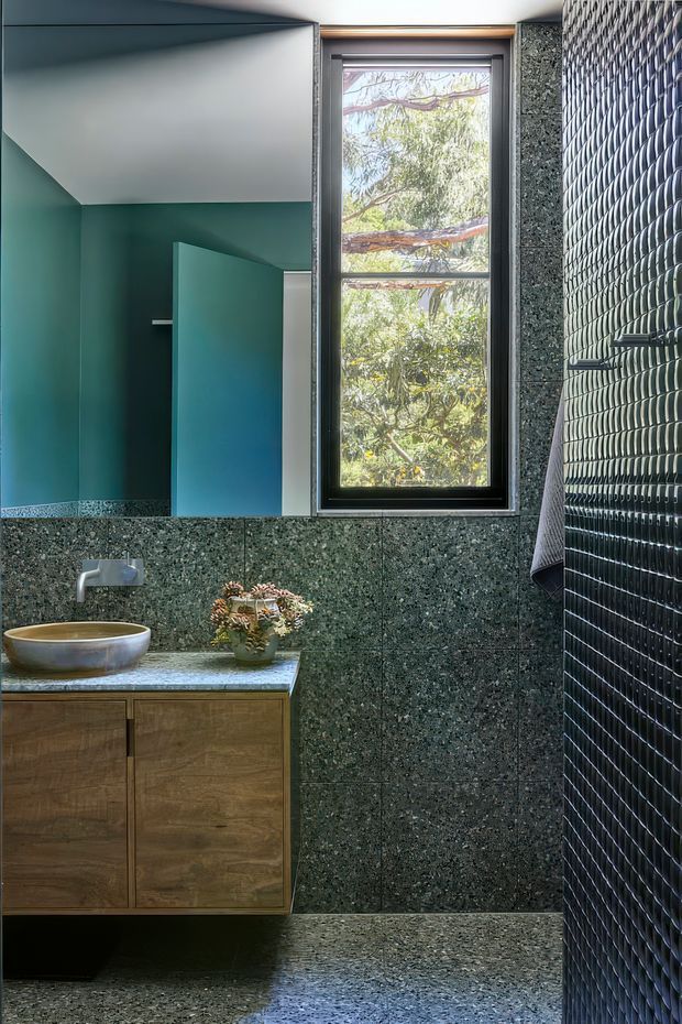 A contemporary bathroom with a floating timber vanity, round vessel basin, wall-mounted tap, and dark green-grey terrazzo surfaces extending across the walls and floor. The room includes a vivid teal painted wall and matching door, a tall black-framed window overlooking trees, and a dark glossy textured tile feature wall enclosing the shower area. The bathroom has a refined, moody yet naturally lit character with strong geometric detailing.