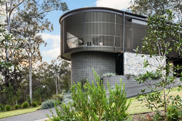 Contemporary curved multi-level building set into a landscaped, wooded site. The facade combines dark vertical louvres forming a screened rounded upper volume, a dark grey glazed-tile cylindrical wall, exposed pale irregular stonework, and dark roof and structural elements. A paved walkway and planted garden surround the building.