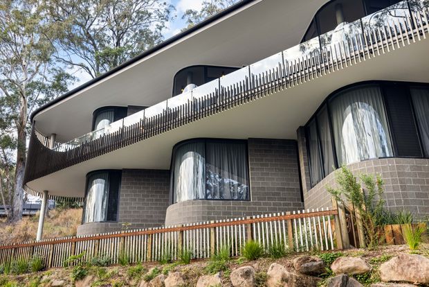 Exterior view of a contemporary multi-level residential building set among trees on a sloping, rocky site. The facade combines dark grey brickwork with smooth light-coloured curved balcony slabs, rounded projecting window bays, extensive dark-framed glazing, and distinctive vertical metal balustrades. Timber-toned fencing and native landscaping appear in the foreground.