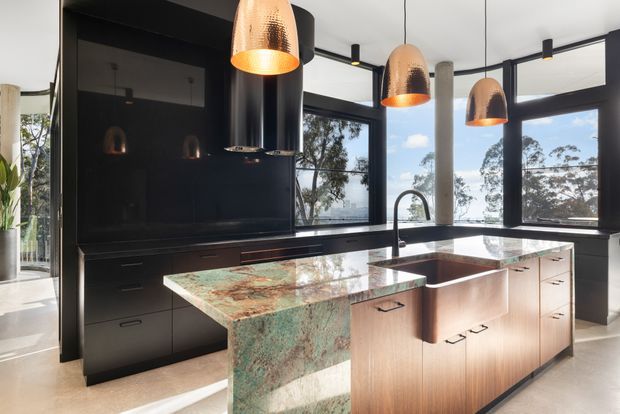 Contemporary kitchen with a large stone island, integrated dark cabinetry, expansive black-framed windows, a sculptural black rangehood, and three copper pendant lights. The island combines a green-and-rust natural stone waterfall end with warm timber cabinetry and an undermount sink. Bright natural light and views of trees outside create a refined, high-contrast interior.
