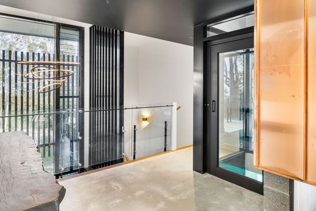 A contemporary interior landing or entry space with a polished concrete floor, dark ceiling, black-framed glazed elevator or doorway, glass balustrade, and tall vertical black metal screening beside floor-to-ceiling glazing. A sculptural pendant light and warm copper-toned wall panel add visual accents. The stairwell or void beyond is visible through the glass railing.