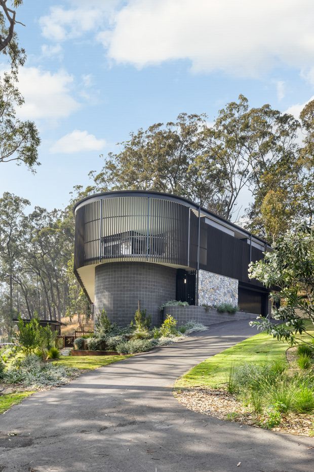 A contemporary dark-clad house set within a wooded, landscaped site. The building has a distinctive curved upper-level facade screened with closely spaced vertical metal fins, a rounded grey masonry base, a projecting cantilevered form, and a sloping roofline. A paved driveway approaches the entrance, with layered native-style planting and mature eucalyptus trees surrounding the home.