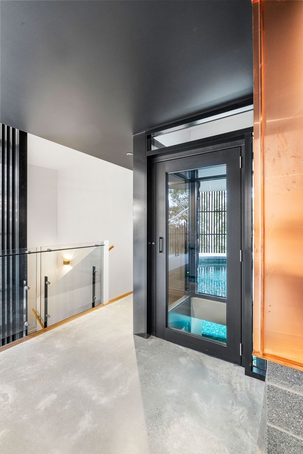 Interior view of a modern upper-level landing or hallway with a black-framed glazed door, glass balustrade, dark ceiling, polished concrete floor, and a glimpse of a turquoise swimming pool through the glazing. An orange-toned exterior wall or cladding panel is visible at the right edge.