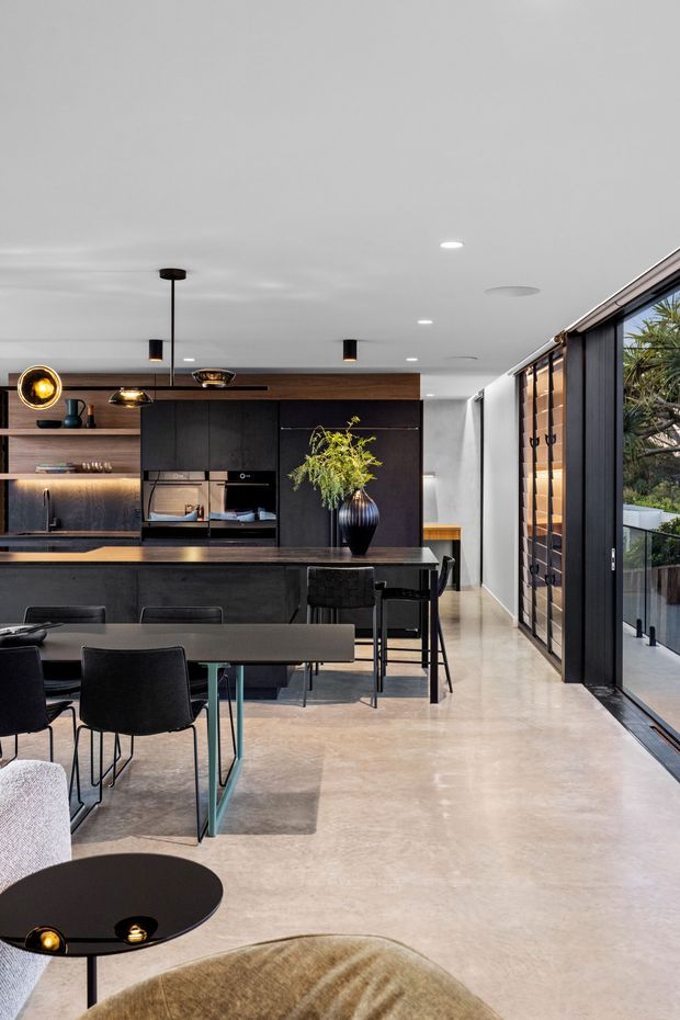 A sophisticated open-plan kitchen and dining area with dark matte cabinetry, integrated appliances, a long island with bar seating, and a dark dining table with black chairs. Warm timber shelving and cabinetry accents contrast with polished concrete flooring, while large glazed sliding doors open to a leafy exterior. Black and brass pendant lighting, concealed strip lighting, and a sculptural vase add refined character.