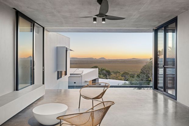 A minimalist modern living area with polished concrete flooring, a smooth concrete ceiling, and expansive floor-to-ceiling glazed openings framing a wide rural landscape and sunset. Sparse sculptural furniture includes woven lounge chairs and a low white round table, while a dark ceiling fan and recessed lighting provide functional accents. The space opens directly onto an elevated outdoor view and appears calm, airy, and highly refined.