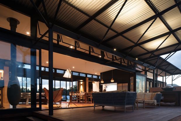 A spacious contemporary indoor-outdoor living area opening onto a timber deck. The room features extensive floor-to-ceiling glazing, exposed black steel framing and trusses, a corrugated metal ceiling, dark built-in cabinetry, pendant lighting, and a lounge and dining arrangement. The covered deck and open structural framework create a strong connection between the interior and exterior.