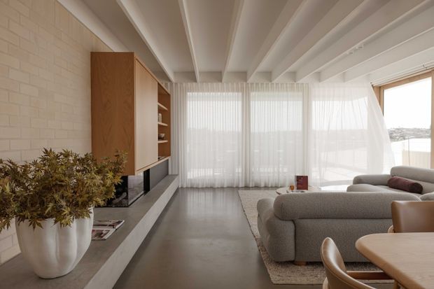 Bright contemporary living room with a sculptural white-painted brick feature wall, built-in timber and neutral-toned joinery, exposed white ceiling beams, full-height sheer curtains, and expansive glazing. A large curved sectional sofa, rug, and dining furniture occupy the open-plan space, with a bright waterfront or coastal outlook visible beyond the windows.