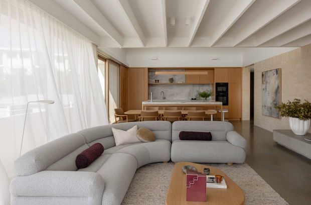 A bright, open-plan contemporary living room with a large curved light-grey sectional sofa in the foreground, opening to a timber-lined kitchen and dining area. The room features full-height sheer curtains, warm timber cabinetry, a pale stone kitchen backsplash and benchtop, polished concrete flooring, a sculptural timber coffee table, and a distinctive white coffered or expressed-beam ceiling. Neutral furnishings and soft natural light create a calm, refined atmosphere.