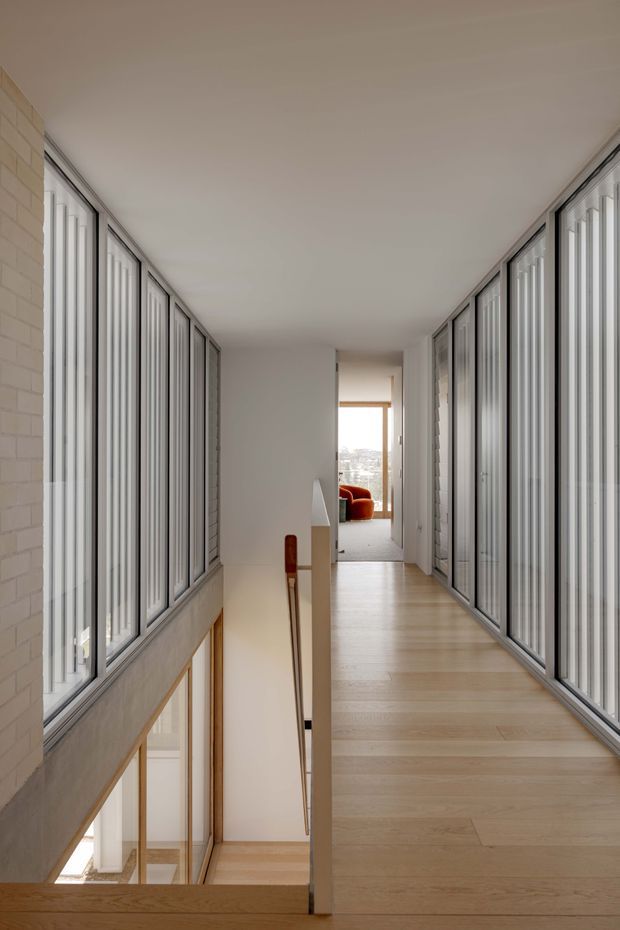 A bright, minimalist upper-level hallway or landing overlooking a stairwell. Full-height glazed panels with slim dark frames line both sides, bringing in abundant daylight, while pale timber flooring and white walls create a clean, airy interior. A glimpse of a vivid orange chair is visible in the room at the end of the corridor.