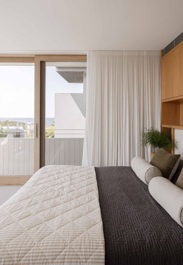 A bright, refined bedroom interior viewed toward a balcony through large sliding glass doors. The room features a neatly made bed with a light quilt and dark textured runner, layered cylindrical cushions, full-height white curtains, pale timber-framed glazing, and built-in warm timber shelving and cabinetry. A coastal view is visible beyond the balcony, while a small indoor plant adds natural character.