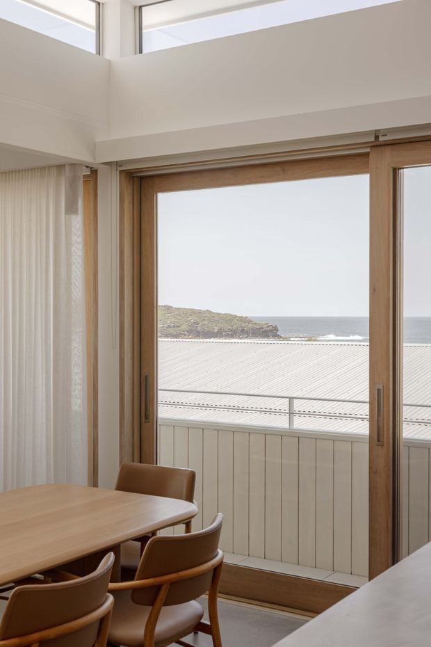 A bright, refined dining area viewed toward a timber-framed glazed door opening onto a balcony or terrace. The room has a pale neutral palette, a timber dining table with leather-upholstered wooden chairs, sheer curtains, and a high ceiling with clerestory glazing. Beyond the balcony, a corrugated light-coloured roof and ocean shoreline are visible.