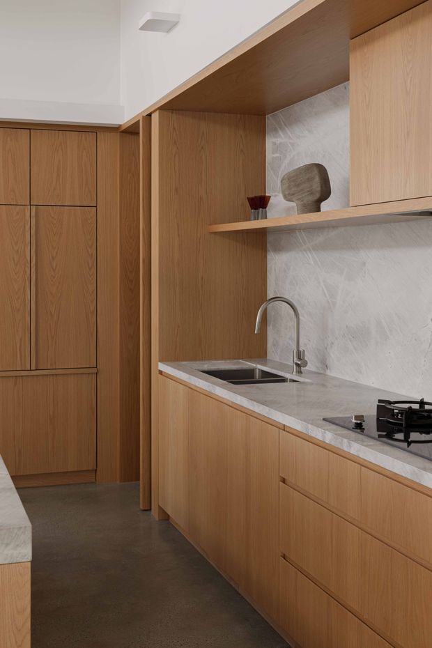 A compact, refined kitchen with extensive warm timber cabinetry, integrated tall storage, a grey stone benchtop, undermount stainless-steel sink and curved tap, gas cooktop, and a pale grey veined stone splashback. Open timber shelving displays a small sculptural object. The space has a clean, contemporary built-in design with a dark polished concrete-looking floor and neutral white walls.