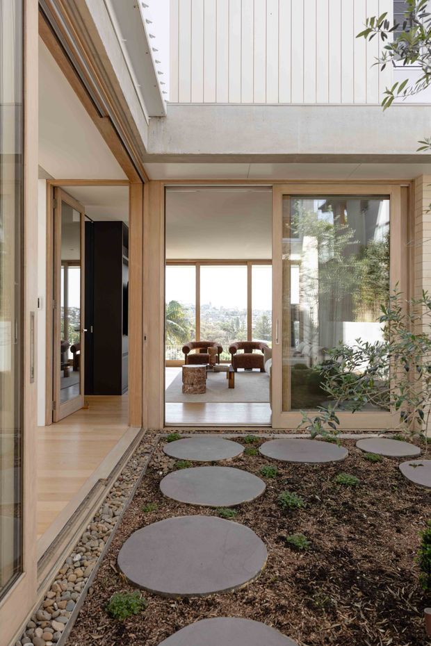 A bright contemporary courtyard viewed from within a home, with circular concrete stepping stones set in a planted garden bed and a pebble drainage strip along the timber-framed glazing. Open sliding doors lead into a refined living room with timber floors, low lounge chairs, and expansive views beyond. The composition highlights indoor-outdoor connection, exposed concrete construction, vertical white cladding, and warm timber detailing.