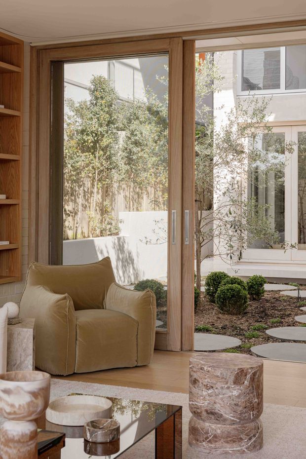A warm, refined living room with full-height timber-framed glazed doors opening onto a landscaped courtyard. The room features a sculptural upholstered armchair, built-in timber shelving, light timber flooring, a low reflective coffee table, and a veined stone side table. Soft neutral tones and abundant natural light create a calm indoor-outdoor atmosphere.